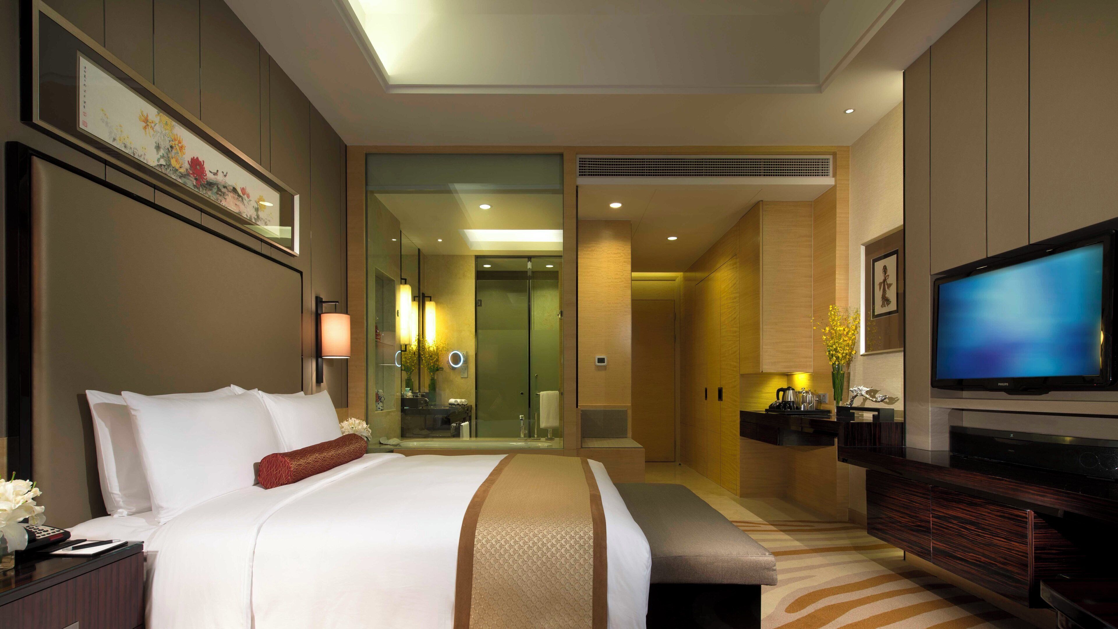 Quarto de hotel com cama, televisão, banheiro e escrivaninha com luminária.