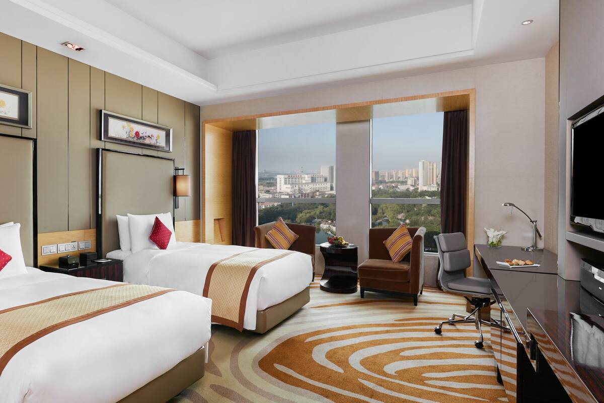 A hotel room with two beds, a sofa, a desk, and a large window offering a city view.