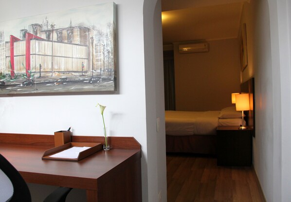A hotel room with a wooden desk, a framed cityscape painting, a bedside table with a lamp, and a bed with white linens. - H3 Hotel Paulista (São Paulo)