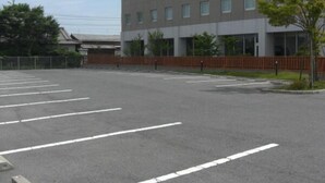 A parking lot with marked parking spaces, a building with multiple windows, and a fenced area with trees. - Hirata MAPLE Hotel (Izumo)