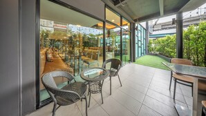 A modern outdoor seating area with glass tables and wicker chairs, a glass wall, and a view of a green space and buildings in the background. - M2 de Bangkok (Bangkok)