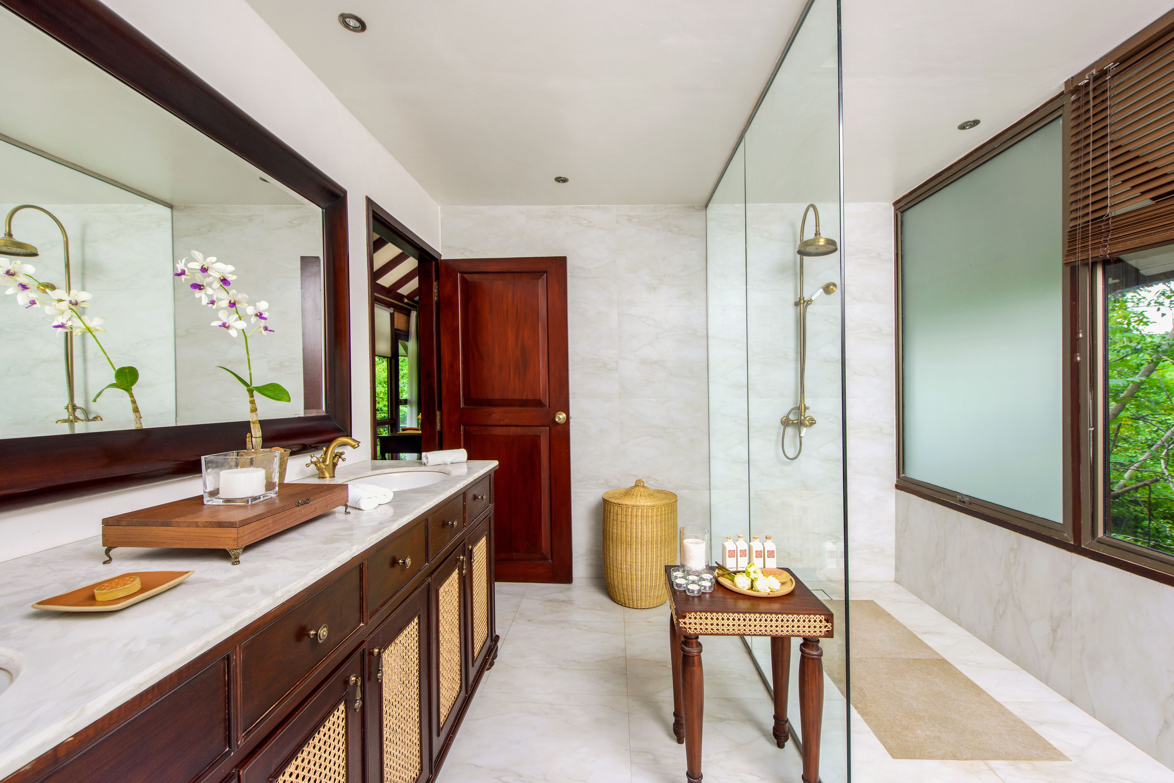 A modern bathroom with a large mirror, marble countertops, a wooden vanity, a glass shower enclosure, and a small table with toiletries.