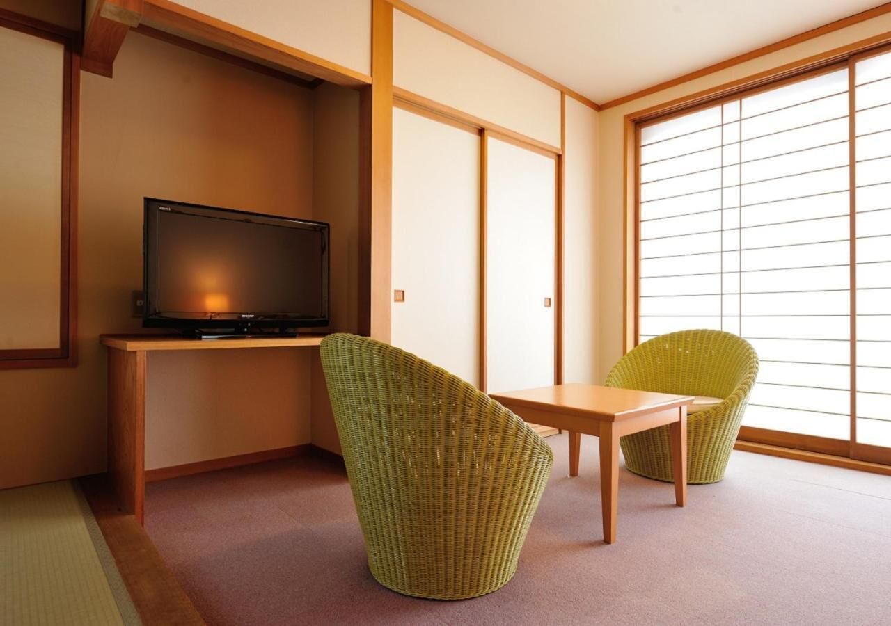 A room with a television on a wooden stand, two green chairs, a small wooden table, and a sliding glass door leading to a bright area.