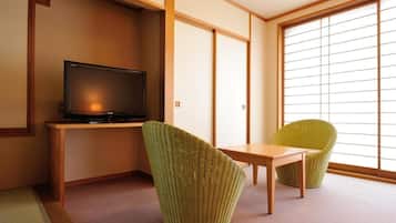 A room with a television on a wooden stand, two green chairs, a small wooden table, and a sliding glass door leading to a bright area.