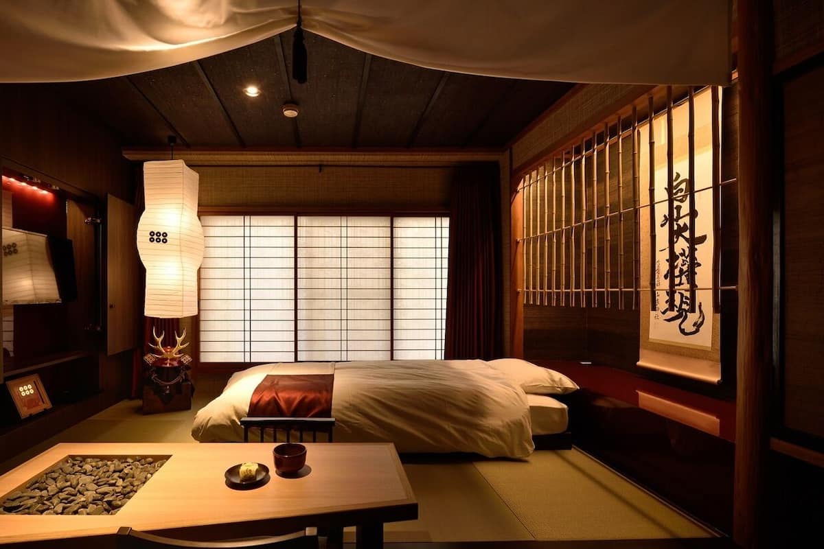 Une pièce traditionnelle japonaise avec un sol recouvert de tatamis, une petite table sur laquelle se trouvent un bol et une assiette, un lit avec une literie blanche, et un paravent shoji orné de calligraphie.
