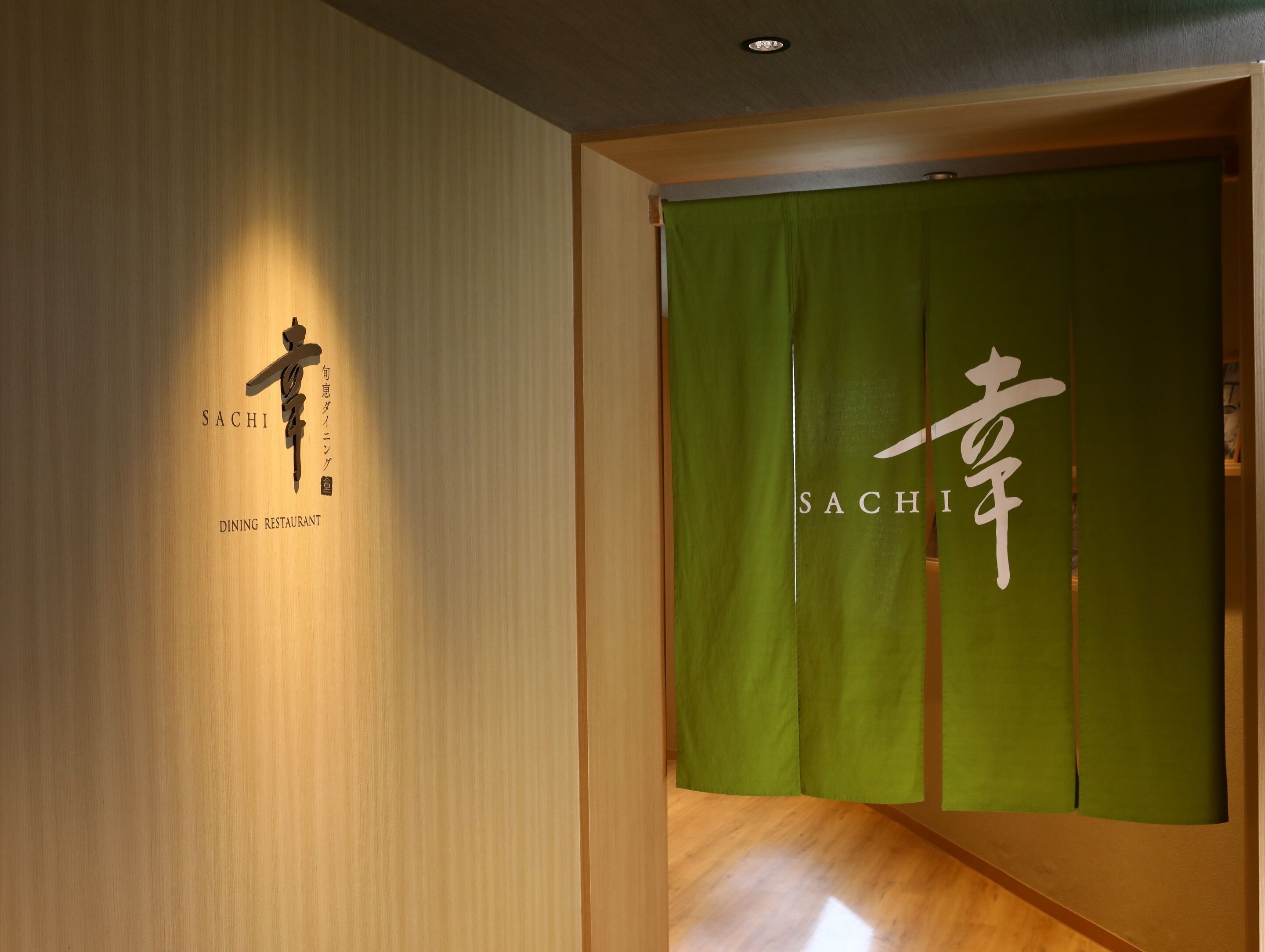 A wooden wall with a sign reading "SACHI DINING RESTAURANT" and a green curtain with Japanese characters.