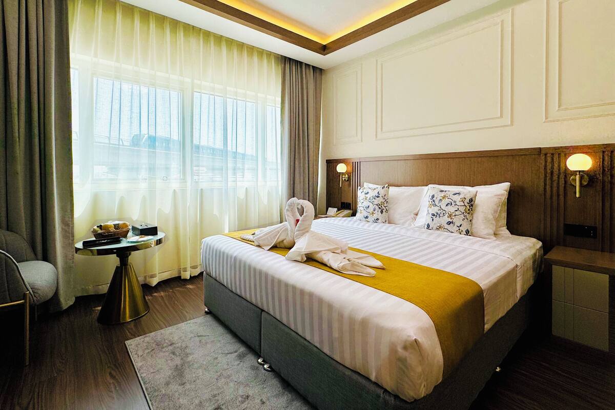 A hotel room with a large bed, a bedside table, a chair, and a view of the city through the window.