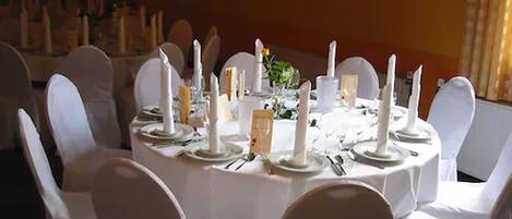 Une table de salle à manger ronde, recouverte de nappes blanches, agrémentée de bougies et de verres, et entourée de chaises blanches.