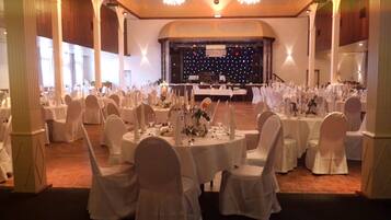Une salle de banquet avec des tables rondes dressées pour un événement formel, agrémentée de nappes blanches, de chaises et de centres de table ornés de bougies.