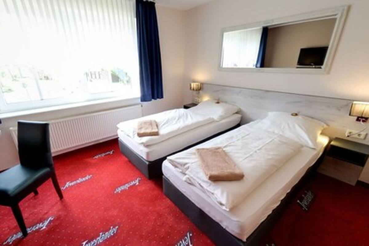 A hotel room with two single beds, a desk, a chair, a window with curtains, and a mirror above the bed.