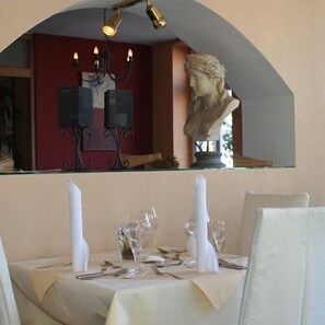 A dining area with a table set for two, a bust sculpture above the table, and a red wall in the background. - Im Schützenhof (Jever)