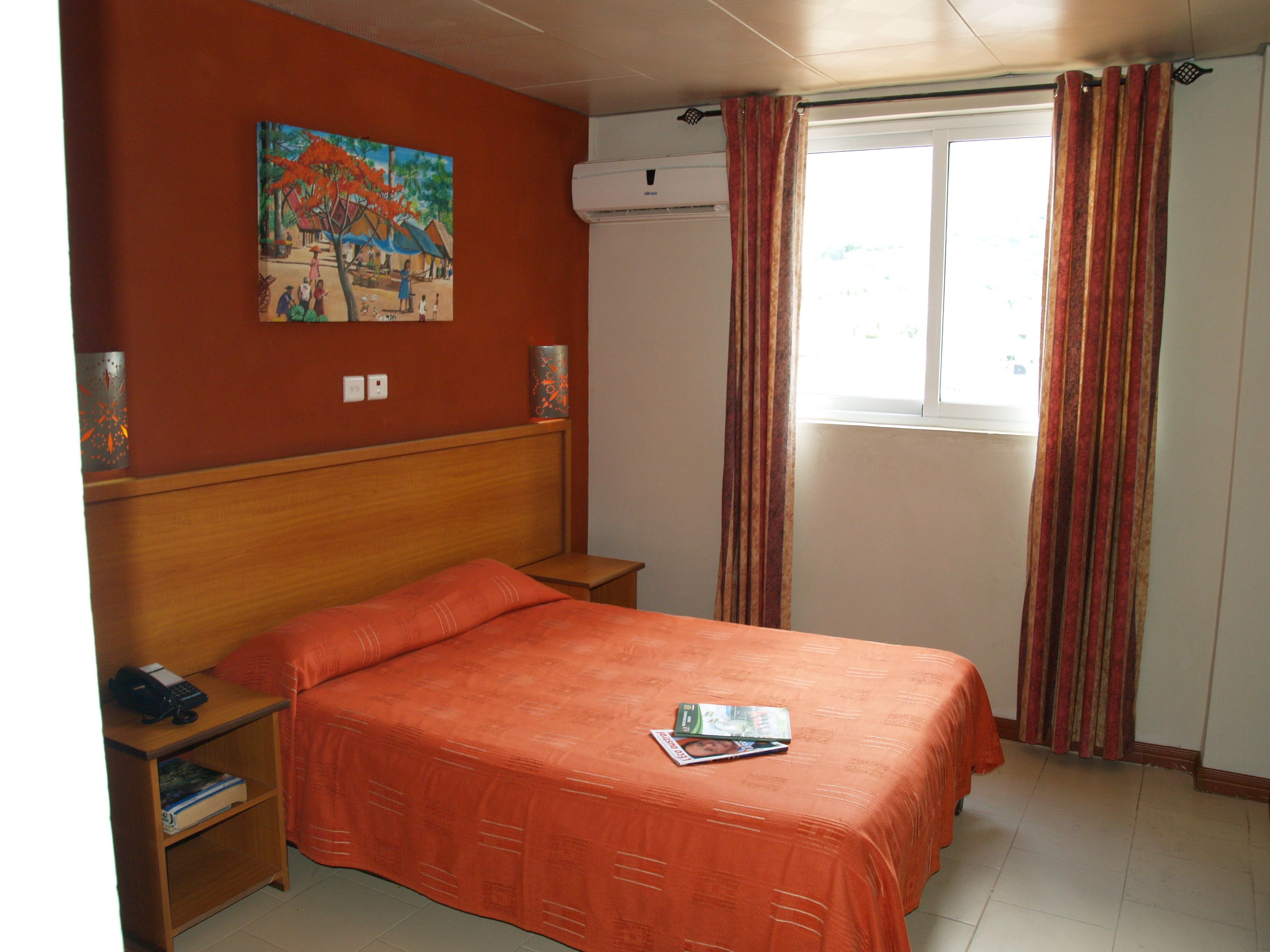 A bedroom with a wooden headboard, a single bed with an orange bedspread, a nightstand with a telephone and books, a wall-mounted air conditioner, and a window with striped curtains.