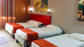 Habitación de hotel con tres camas, cada una con ropa blanca y mantas color naranja. Tiene un cabecero rojo, un cuadro en la pared y una ventana con cortinas.