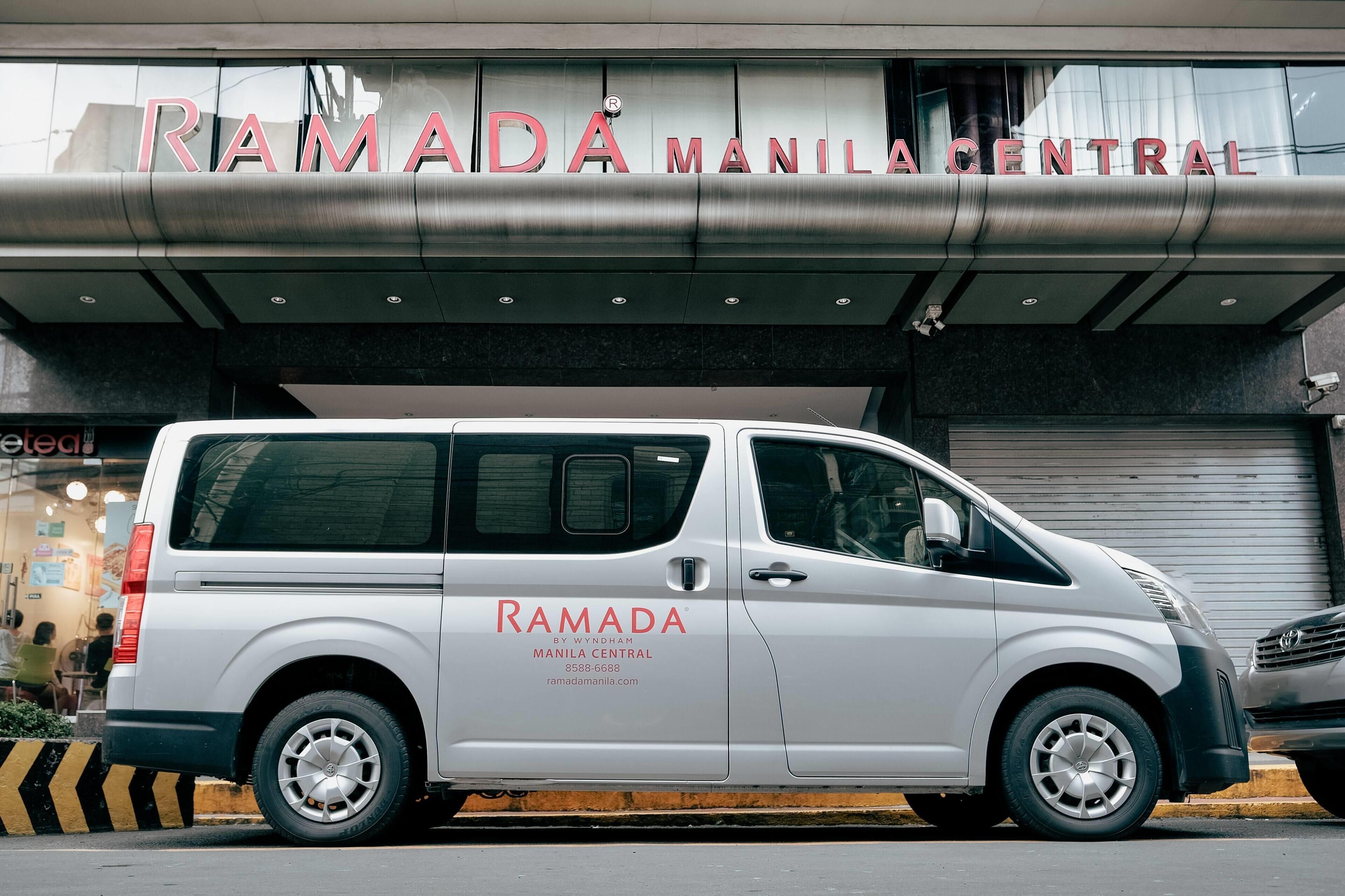 Una furgoneta blanca aparcada davant d'un edifici amb un cartell que diu 'RAMADA MANILA CENTRAL'.