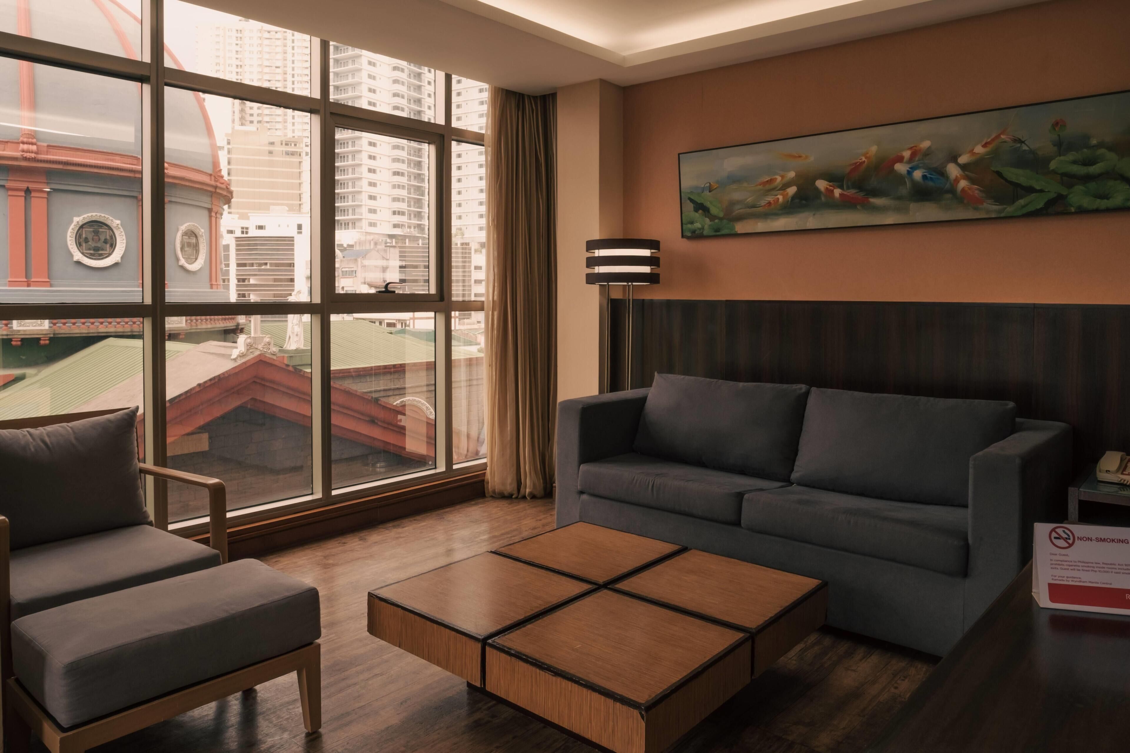 Una habitación de hotel moderna con un ventanal grande, un sofá gris, una mesa de centro de madera y un cuadro de carpas.