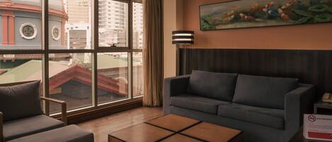 Una habitación de hotel moderna con un ventanal grande, un sofá gris, una mesa de centro de madera y un cuadro de carpas.