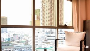 A hotel room with a large window offering a city view, a chair with a cushion, and a wooden nightstand. - Ramada by Wyndham Manila Central (Manila)