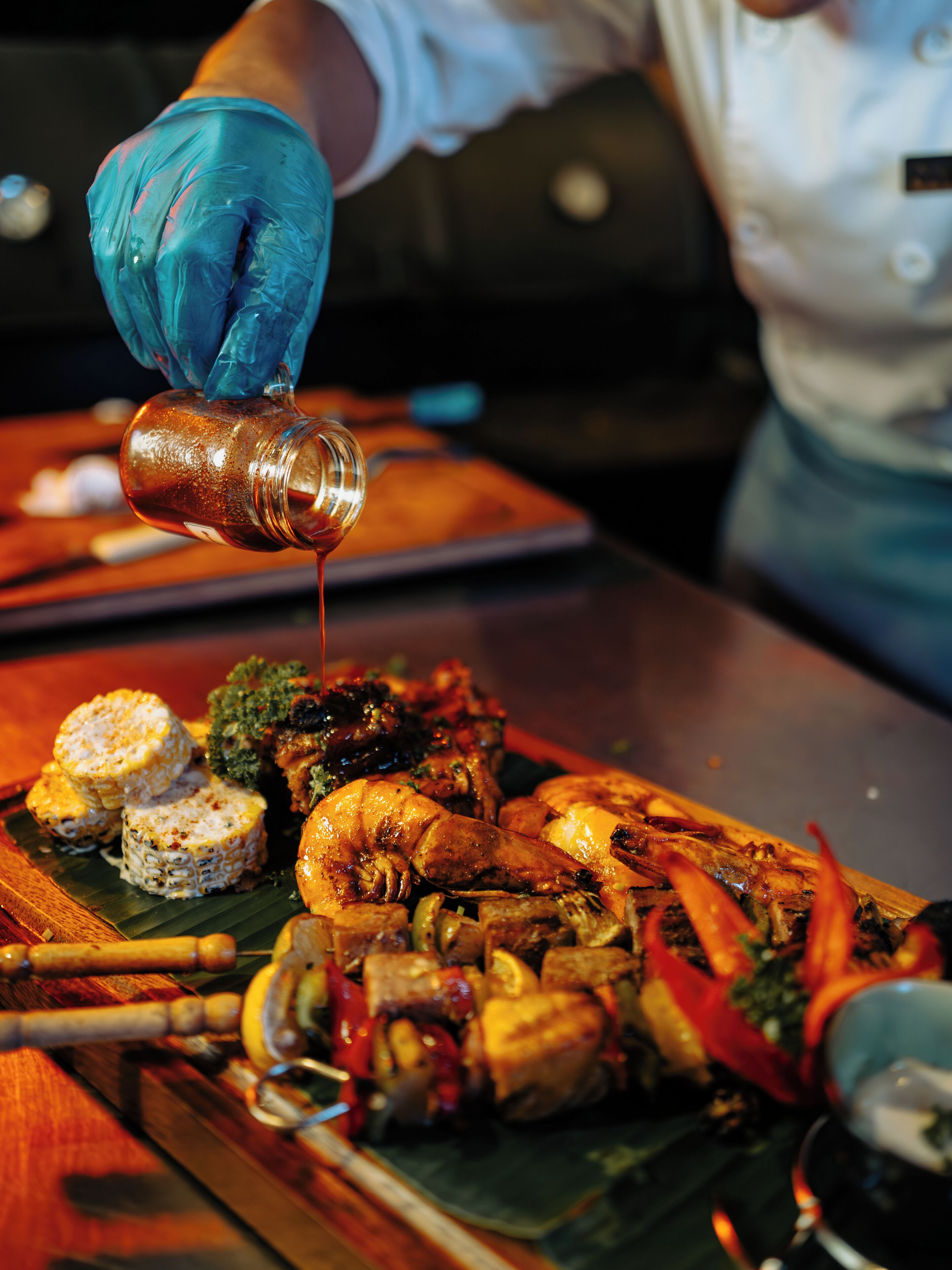A chef pouring sauce over a dish of grilled meats and vegetables.