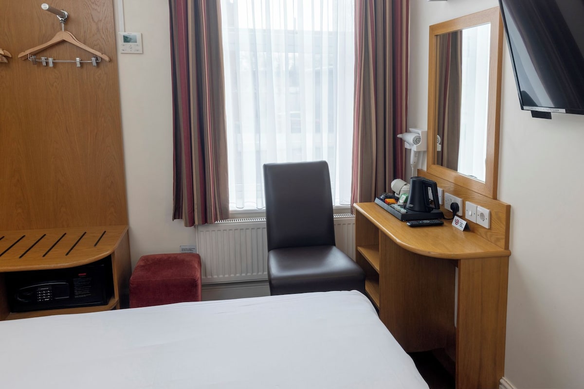 A hotel room with a bed, a desk, a chair, a radiator, a TV, and a window with curtains.