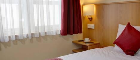 Quarto de hotel com uma cama, mesa de cabeceira, abajur, xícara e controle remoto.