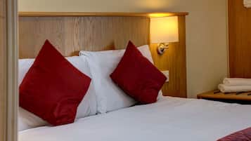A neatly made bed with white linens and two red pillows, a wooden headboard, a bedside lamp, and a wooden nightstand with a folded white towel.