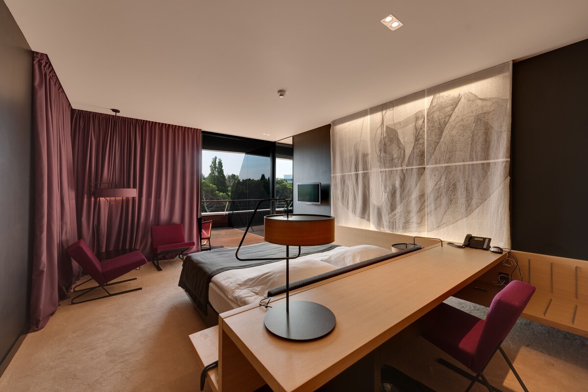 Habitación de hotel moderna con cama, escritorio, sillas, televisor y ventana grande con vistas.