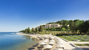 Un resort frente a la playa, con un mar azul cristalino, una playa de arena con sombrillas y un edificio con varios balcones con vista al mar.