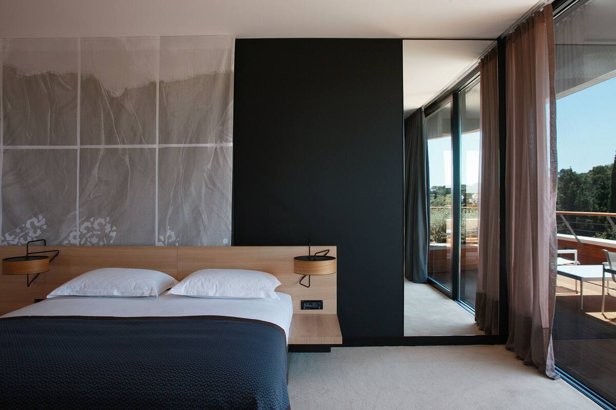 Un dormitorio moderno con una cama grande, una mesita de noche y vistas al exterior a través de un ventanal corredero.