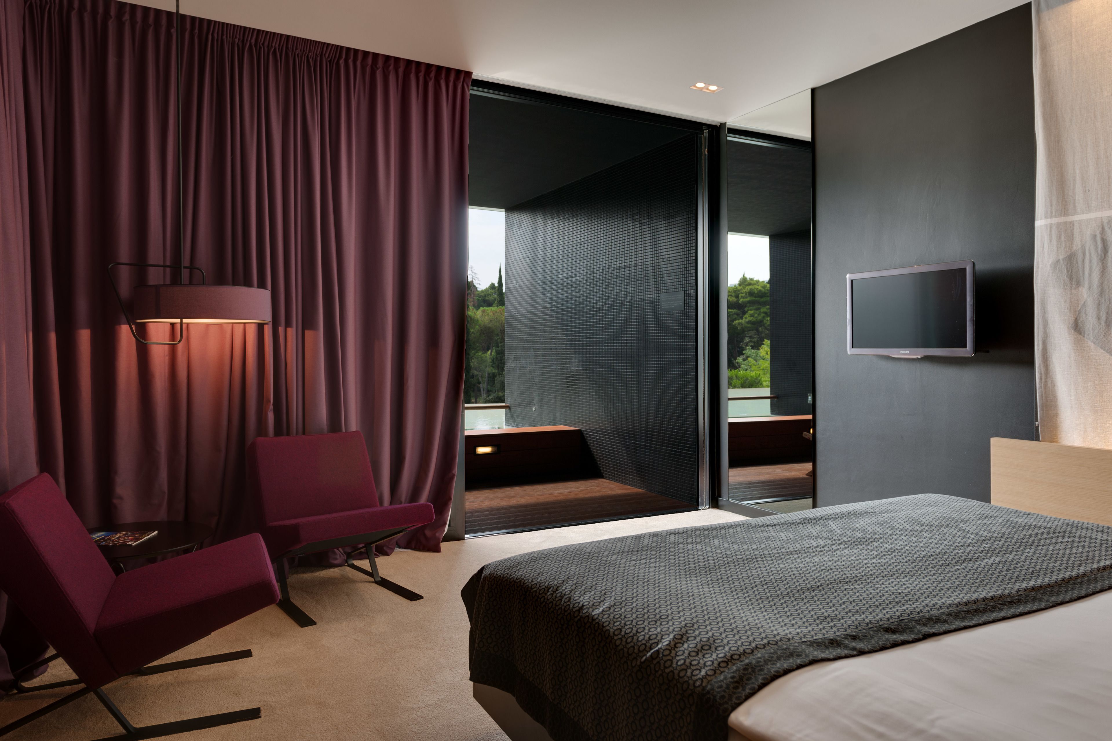 Ett modernt hotellrum med en stor säng, en sittgrupp med en stol och ett bord, en platt-TV som är monterad på väggen och utsikt över grönområden genom glasdörren.