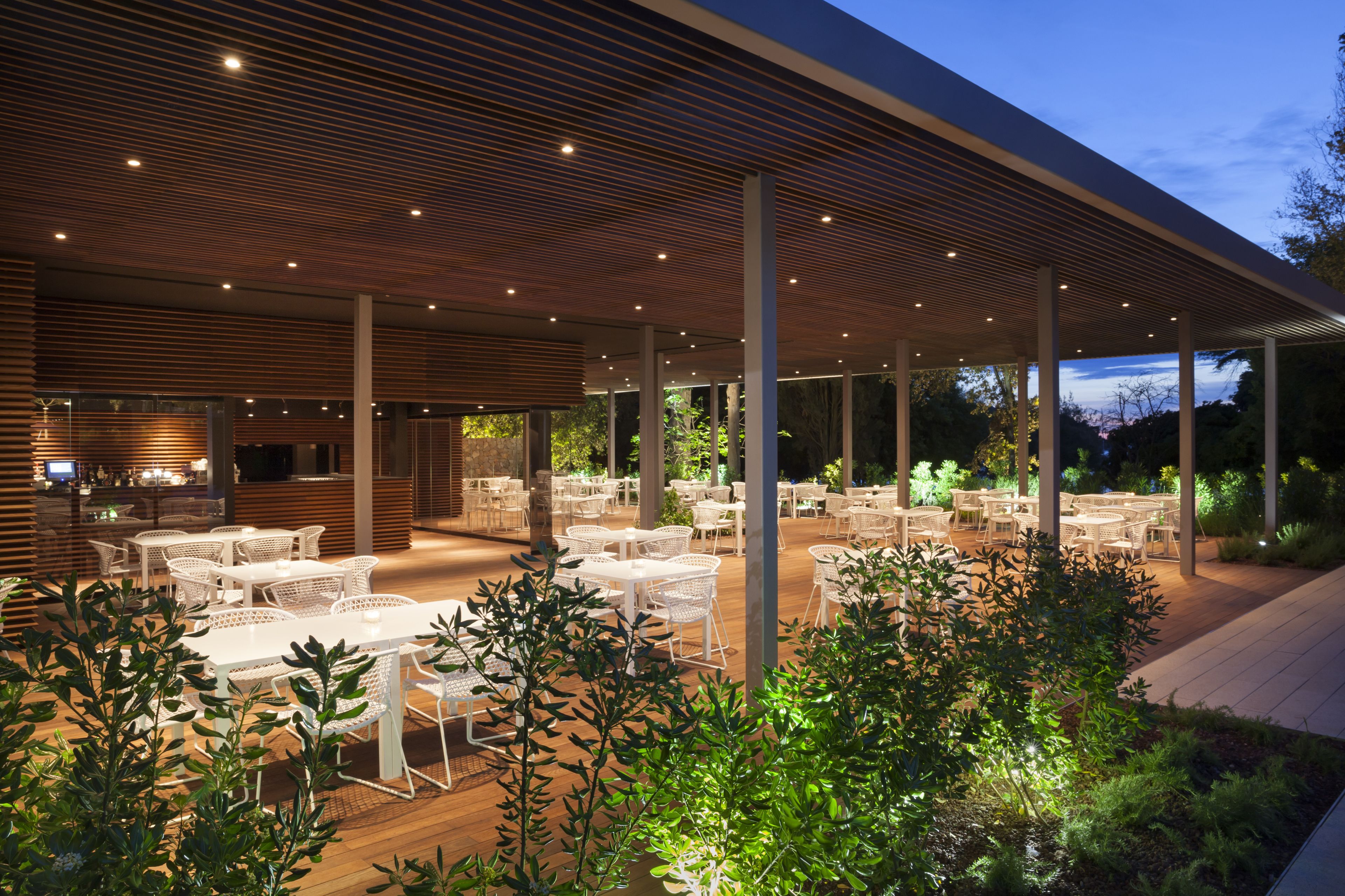 An outdoor dining area with white tables and chairs, wooden slatted ceiling, and greenery.