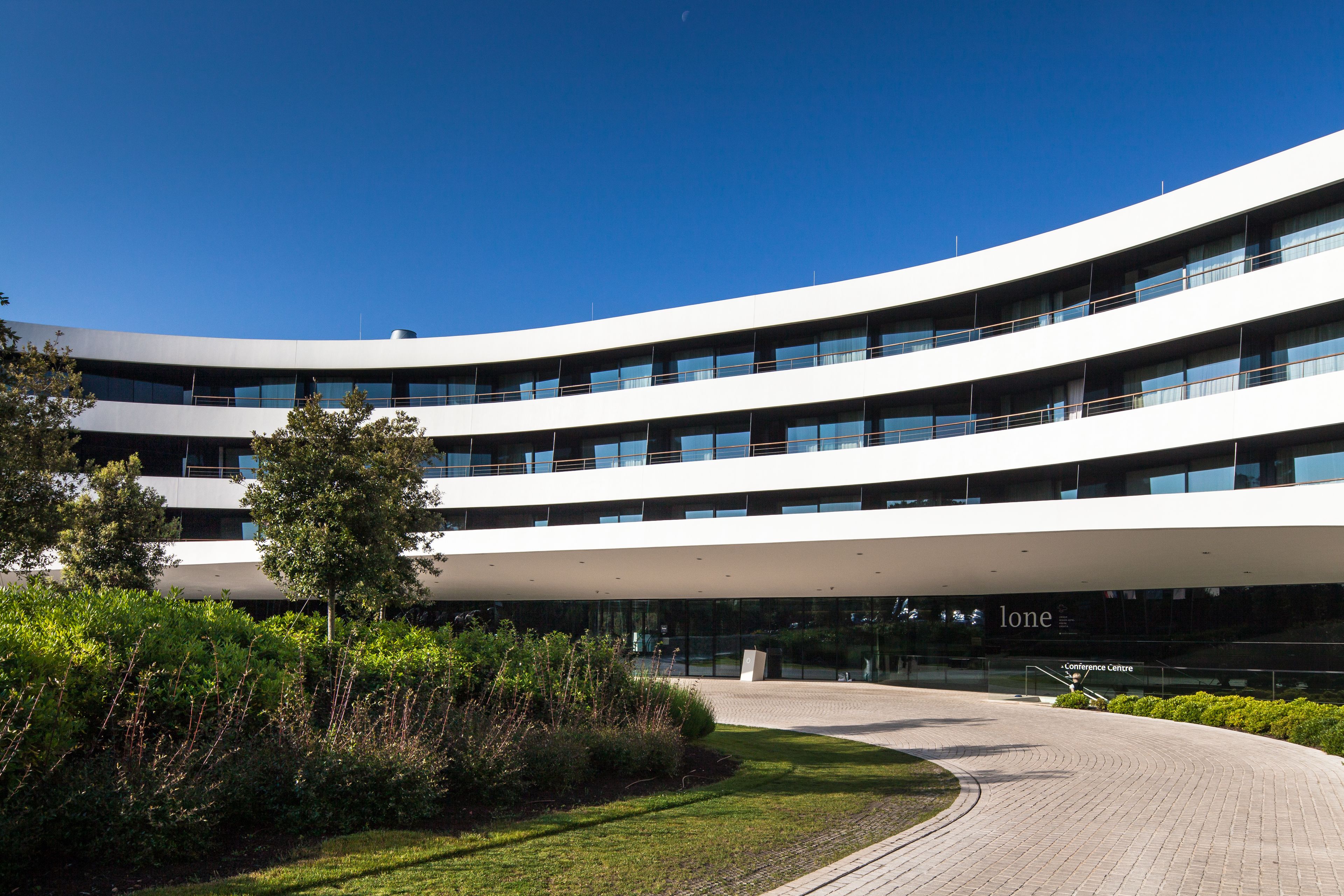 A modern building with a curved facade, multiple balconies, and a landscaped entrance area.