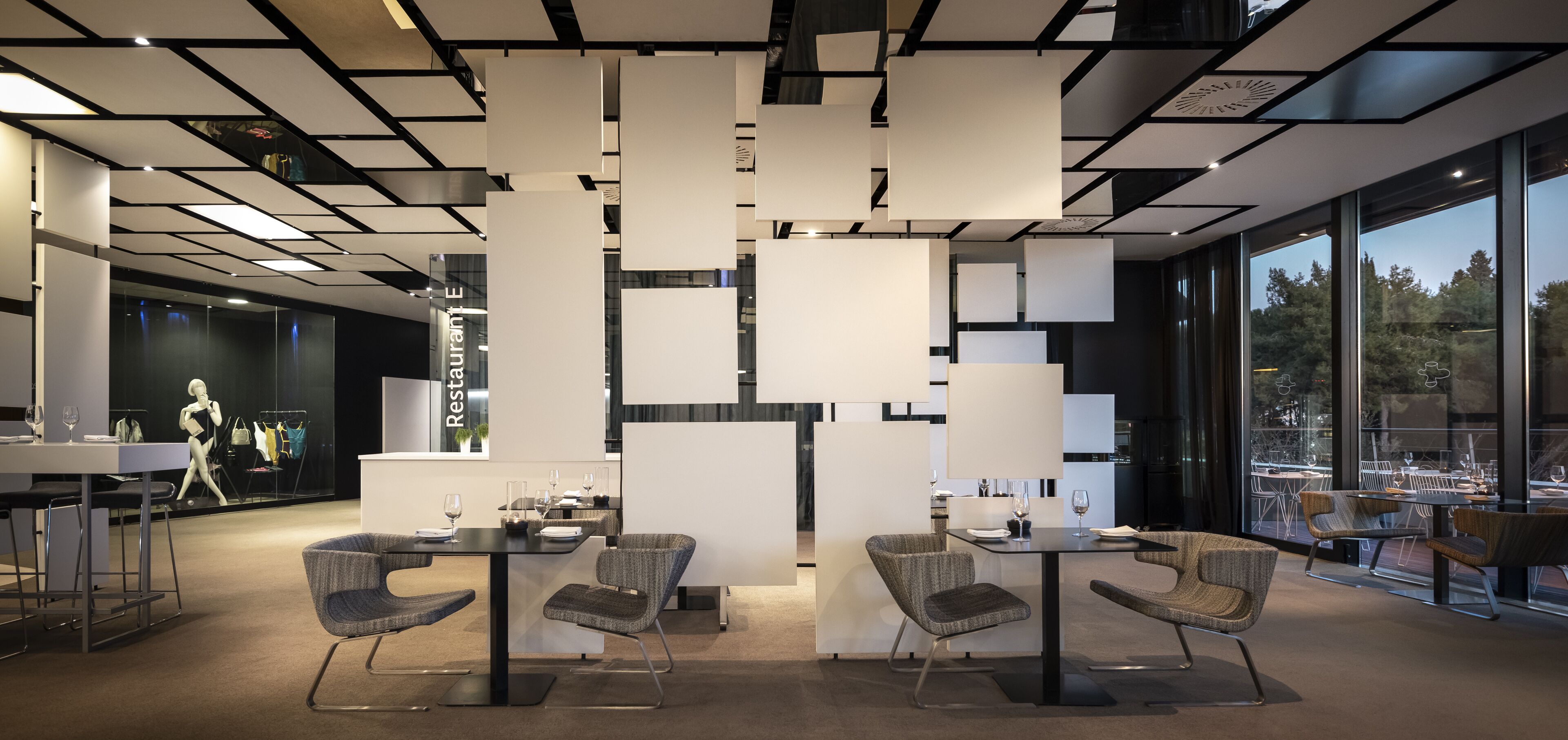 A modern restaurant interior with high ceilings, geometric ceiling panels, and large windows offering a view of the outdoors.