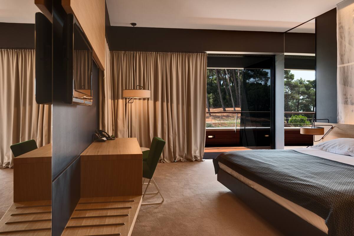 Habitación de hotel moderna con cama, escritorio, silla, lámpara y un ventanal con vistas a árboles.