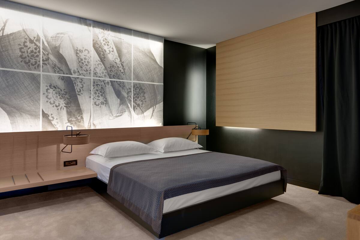 Un dormitorio moderno con una cama grande, una mesita de noche, una lámpara de pared y una ventana con vistas a un edificio.