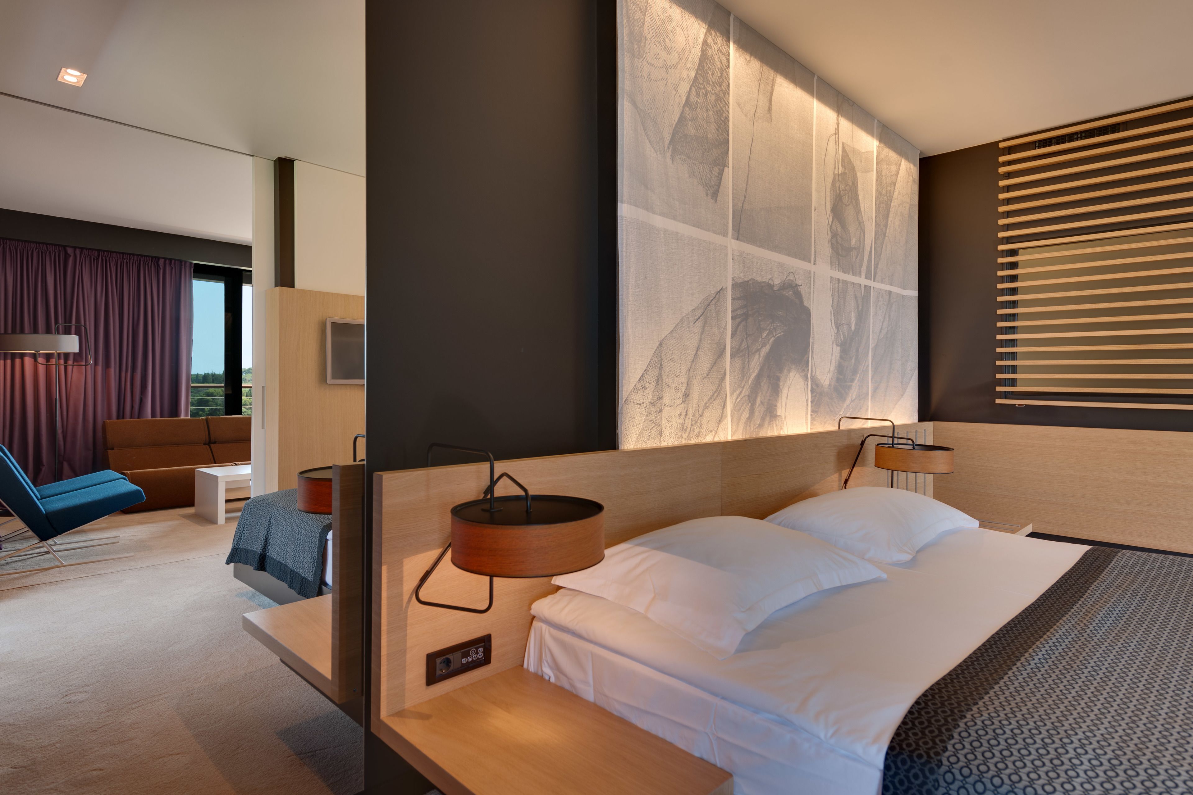 A modern hotel room with a large bed, a bedside table, a wall-mounted TV, and a view of the outdoors through large windows.