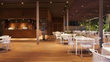 An indoor dining area with white wicker chairs and tables, a bar area with bottles, and a stone wall in the background.