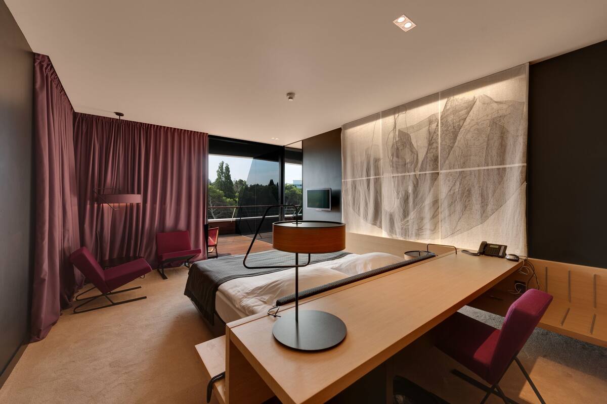 Una habitación de hotel moderna con una cama grande, un escritorio, una silla, un televisor y vistas al exterior.