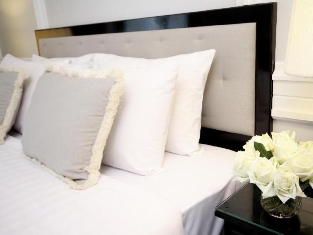 A bed with white bedding and fluffy pillows, a glass side table with a vase of white flowers, and a black headboard.