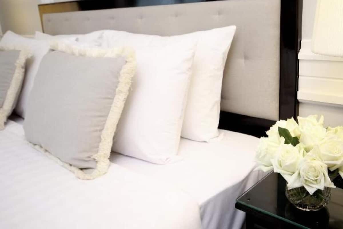 Een bed met witte beddengoed en pluizige kussens, een bijzettafeltje van glas met een vaas witte bloemen, en een zwart hoofdbord.