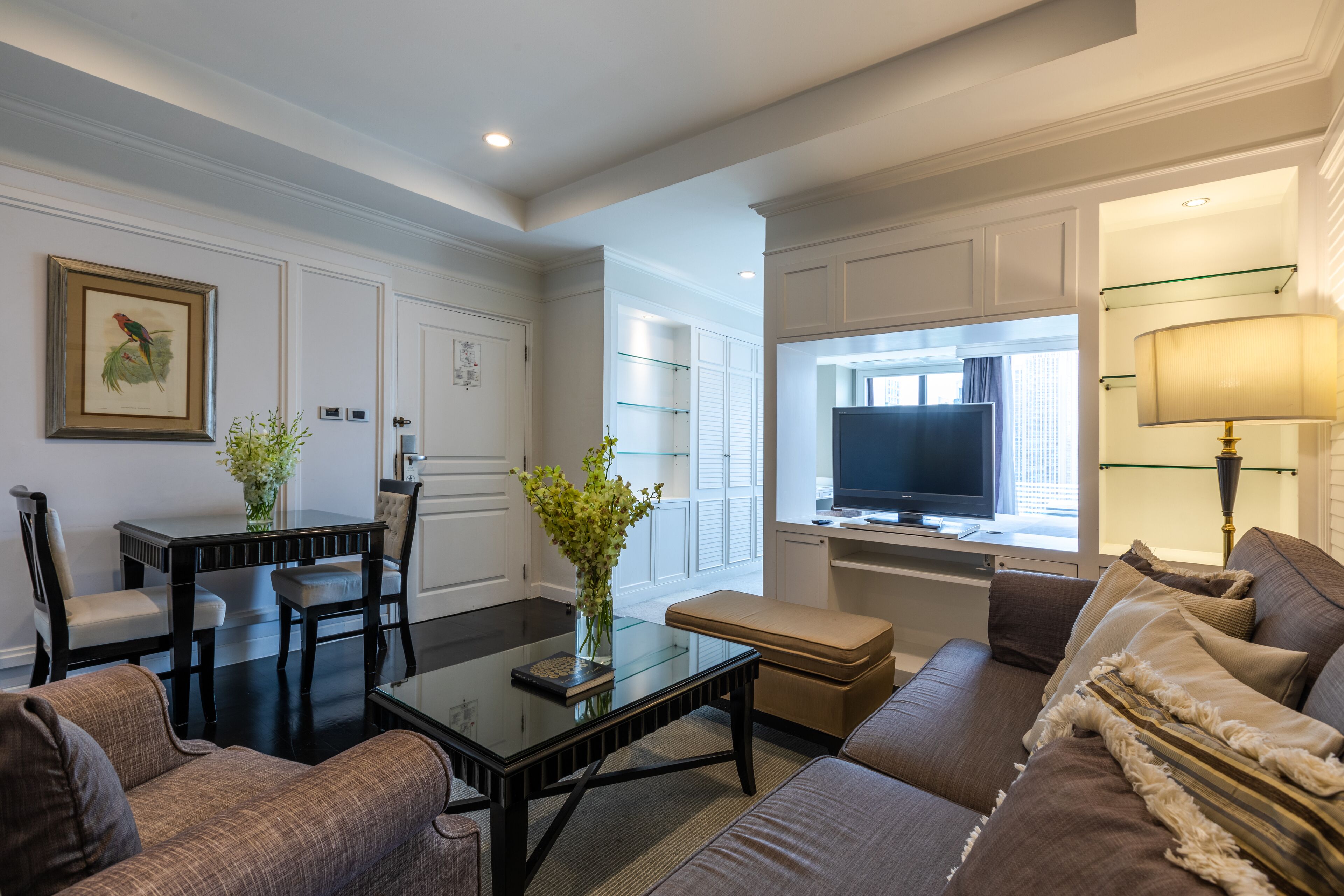 A modern living room with a glass coffee table, a sofa, a TV, and a dining area with chairs and a vase of flowers.