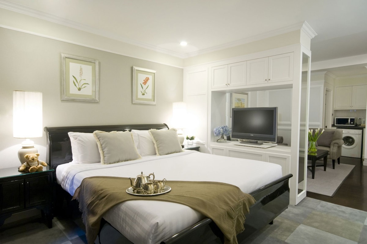 A hotel room with a bed, bedside table with a lamp and teddy bear, a TV, and a kitchenette area.