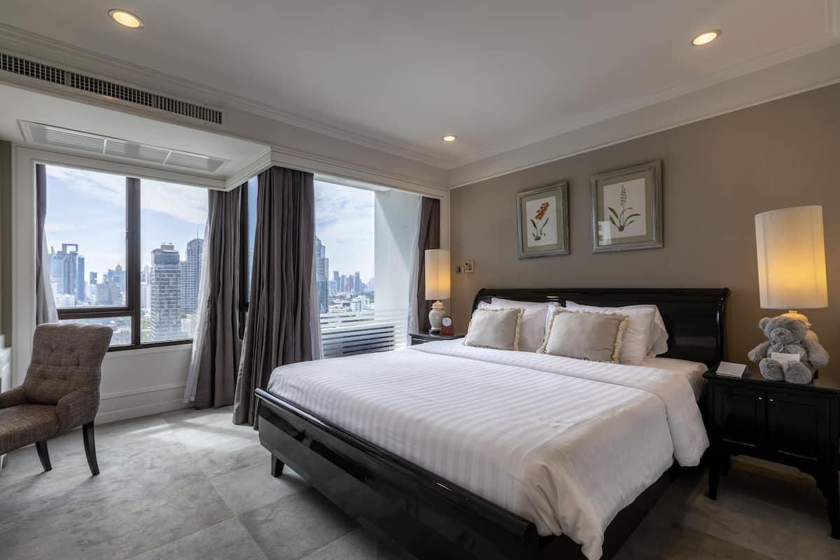 Hotelkamer met een groot bed, een stoel, een nachtkastje met een lamp en uitzicht op de skyline van de stad.
