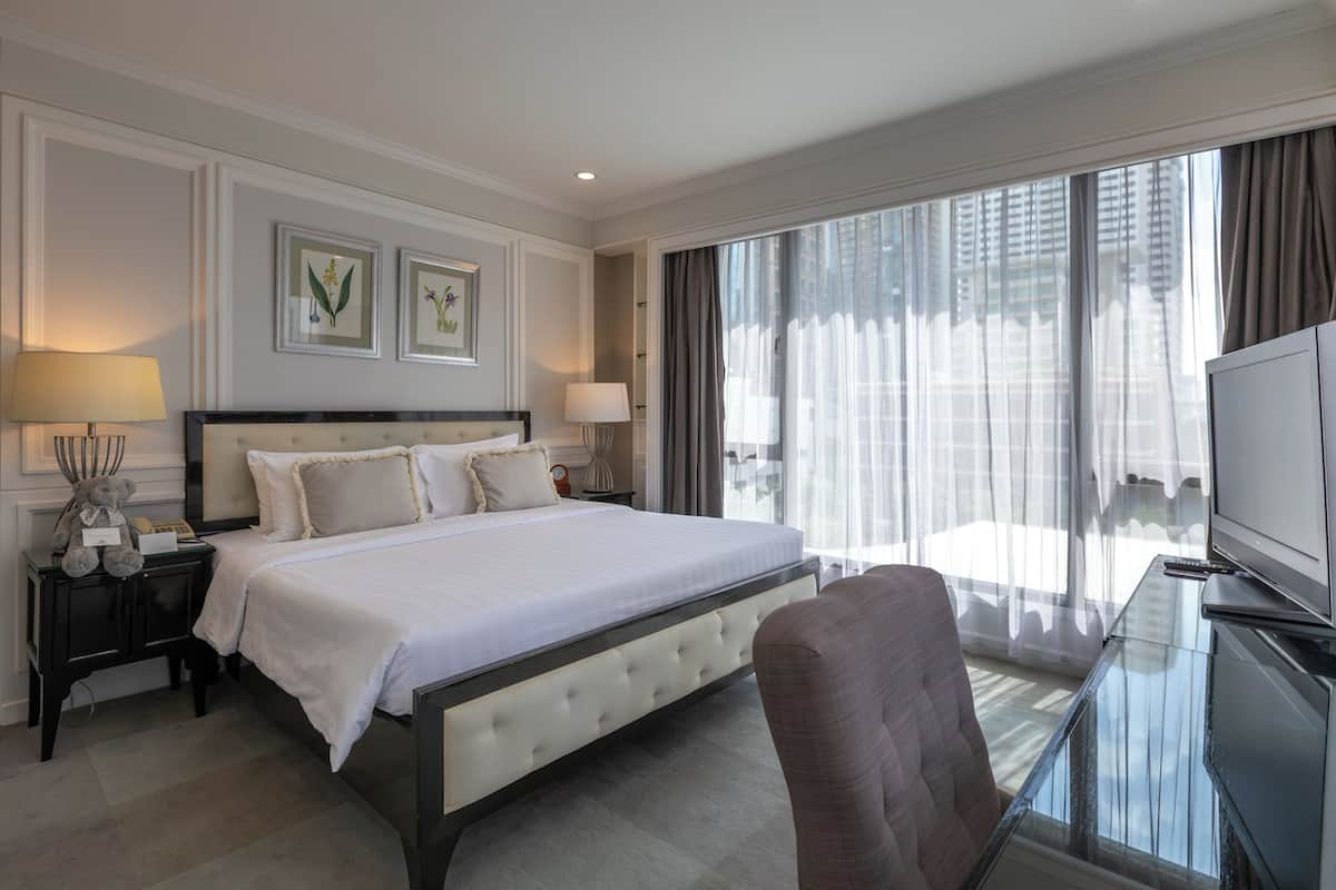 Hotelkamer met een groot bed, een glazen bureau met een televisie en uitzicht op een stadsgezicht door dunne gordijnen.