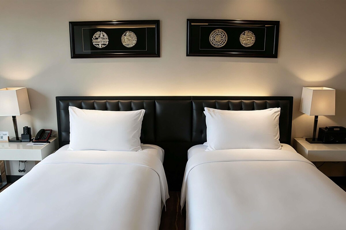 Habitación de hotel con dos camas, cabecero negro, mesitas de noche con lámparas y cuadros decorativos en la pared.