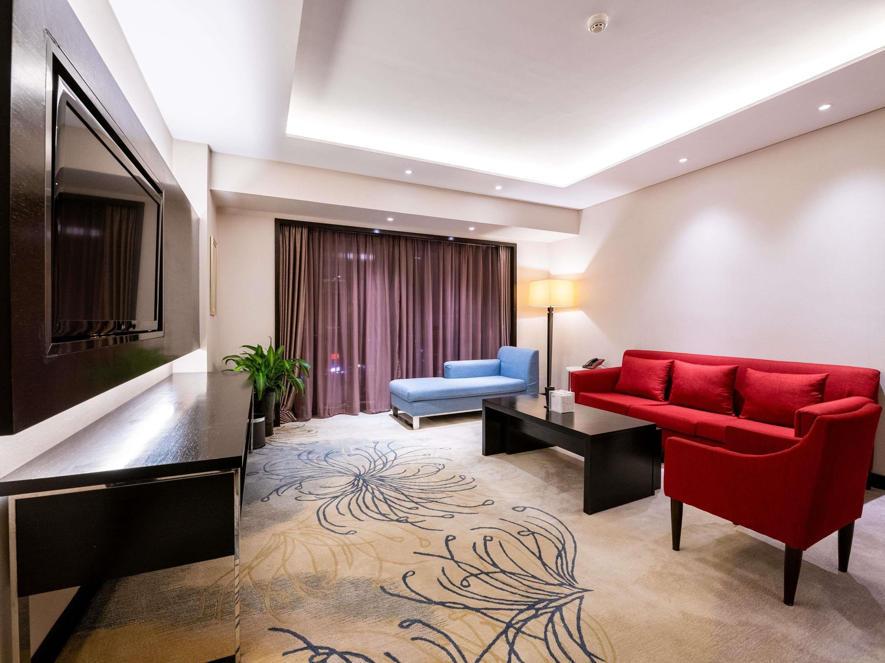 A modern hotel room with a flat-screen TV, a red sofa, a blue sofa, a coffee table, a lamp, and a decorative rug.