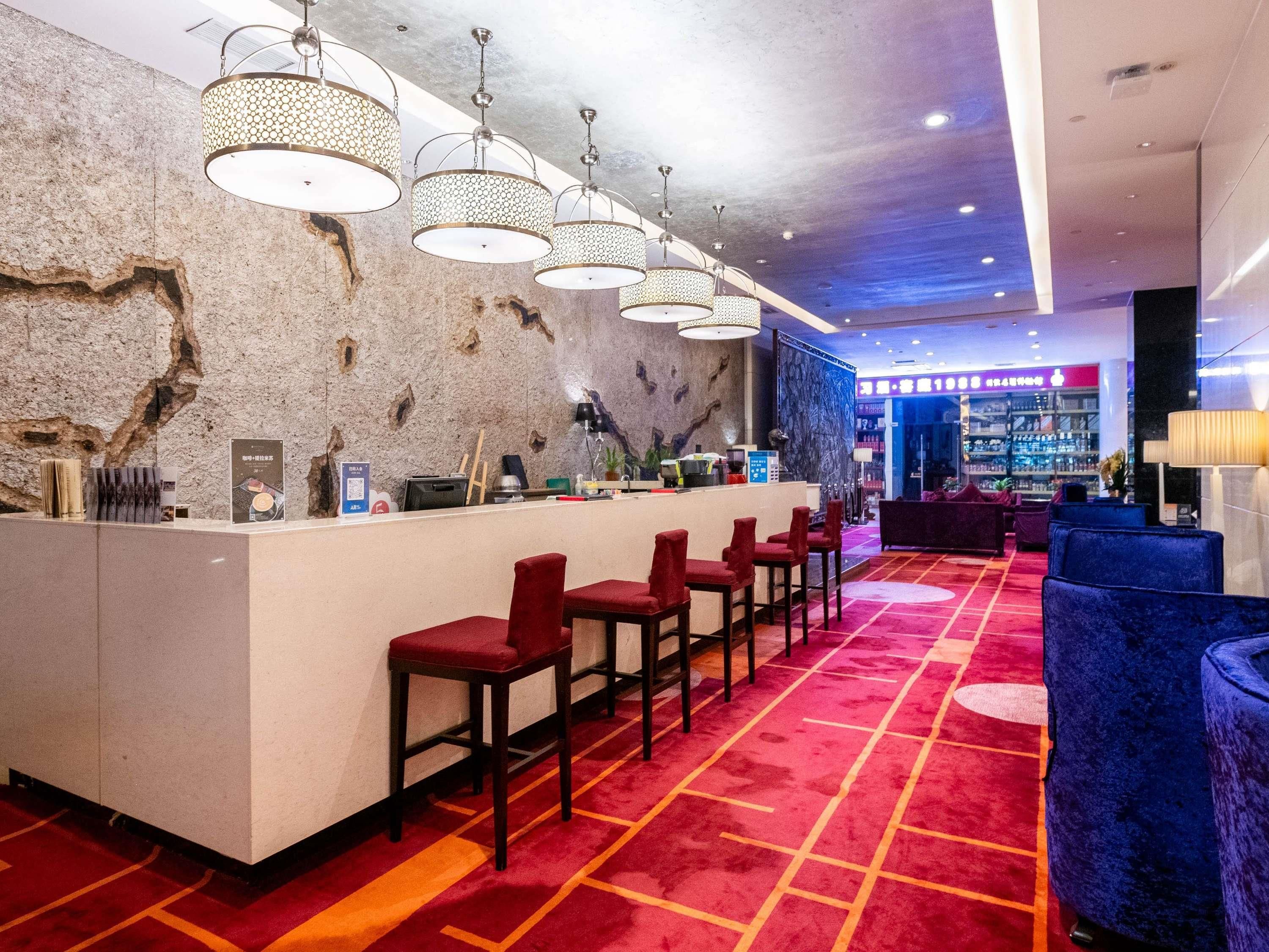Un hall d’hôtel avec un comptoir d’accueil, des tabourets de bar rouges, un mur à motifs et un espace salon bleu.