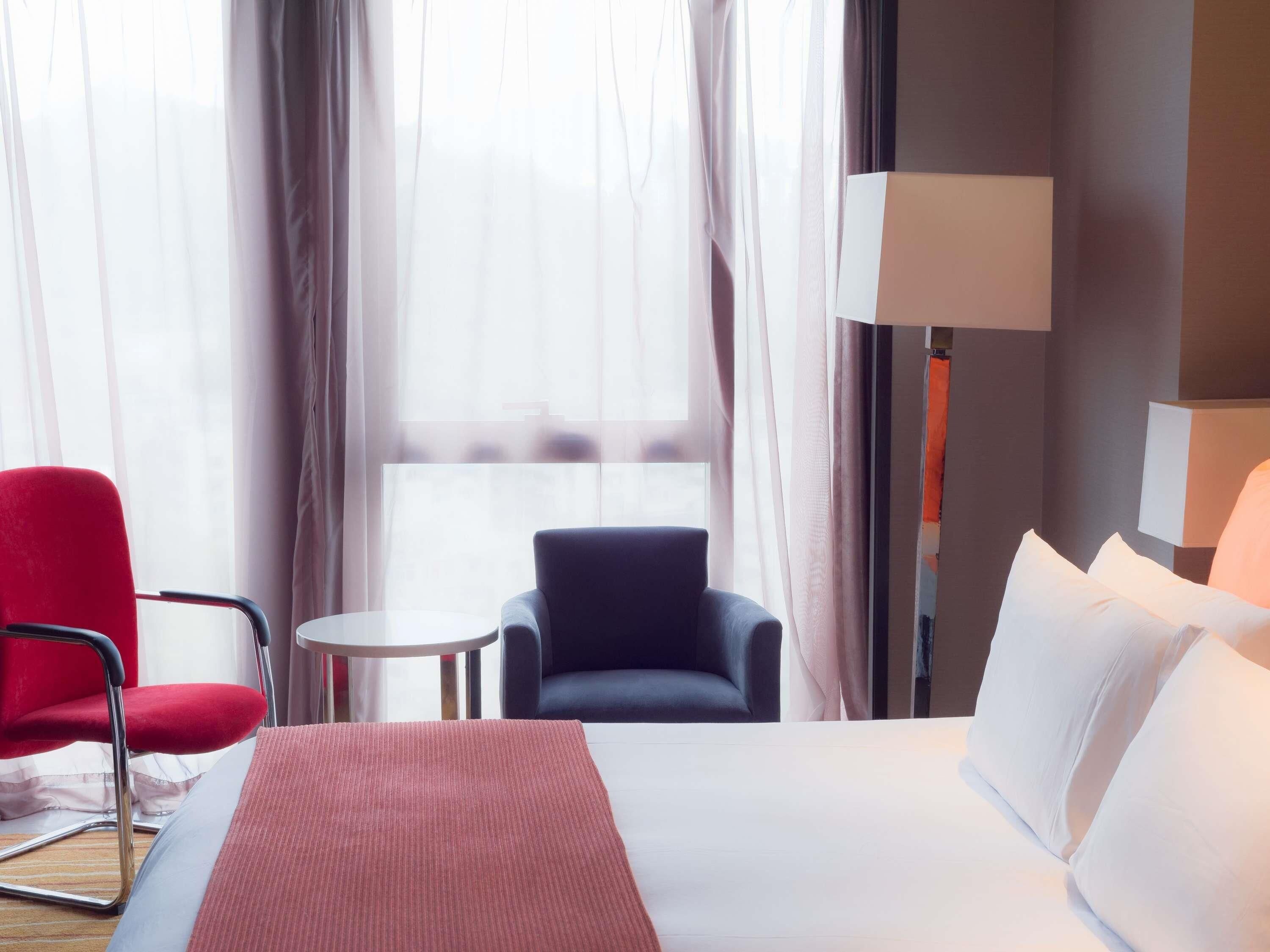 Ein Hotelzimmer mit einem Bett, einem roten Sessel, einem blauen Stuhl, einem kleinen Tisch und einer Lampe.