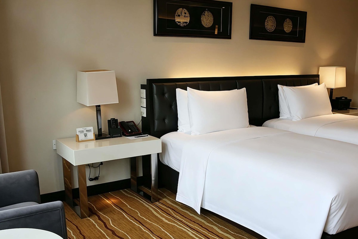 Quarto de hotel com duas camas, uma escrivaninha com telefone, uma cadeira e piso coberto por carpete.