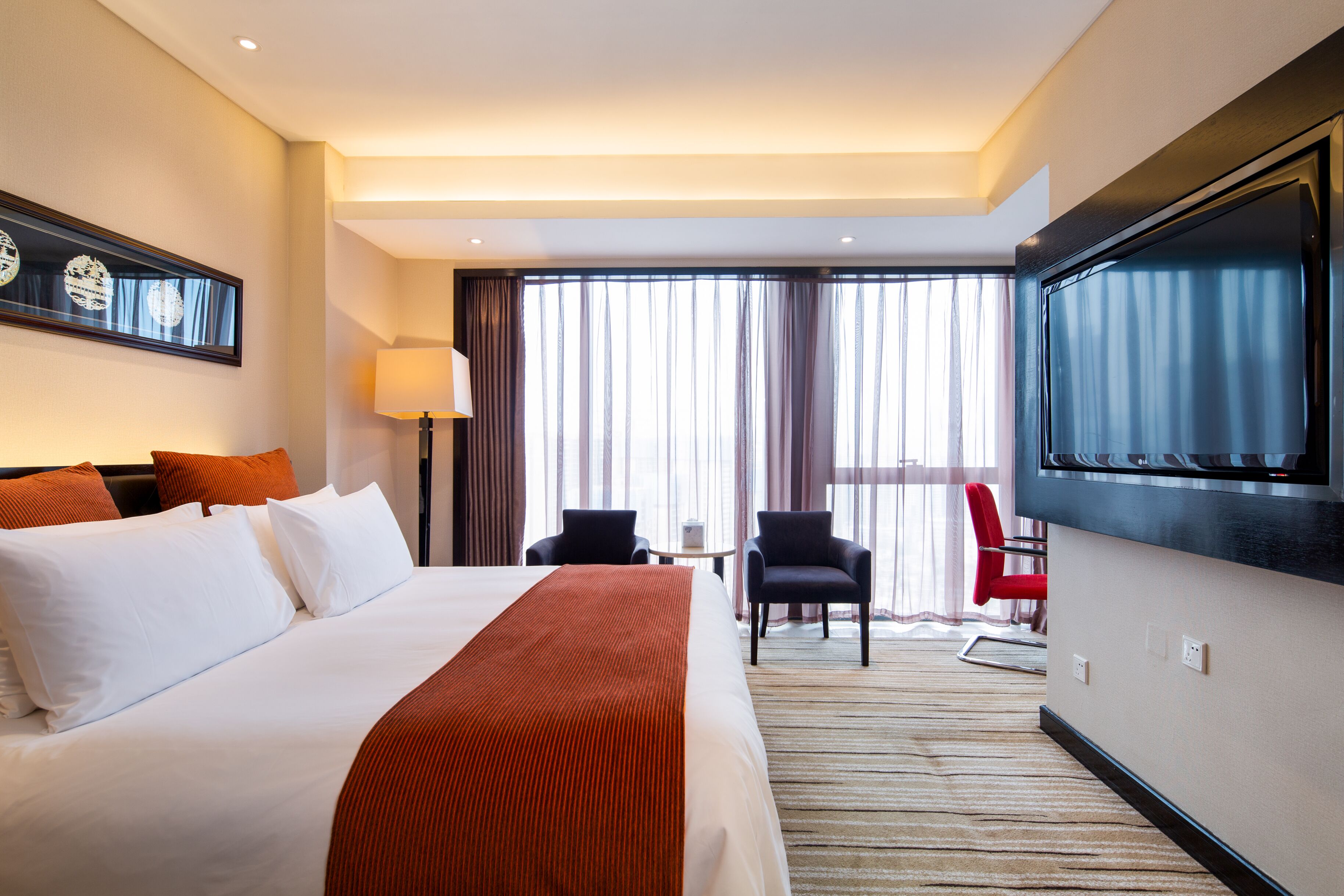 A hotel room with a large bed, a TV mounted on the wall, a desk with two chairs, and a view of the outdoors through large windows.
