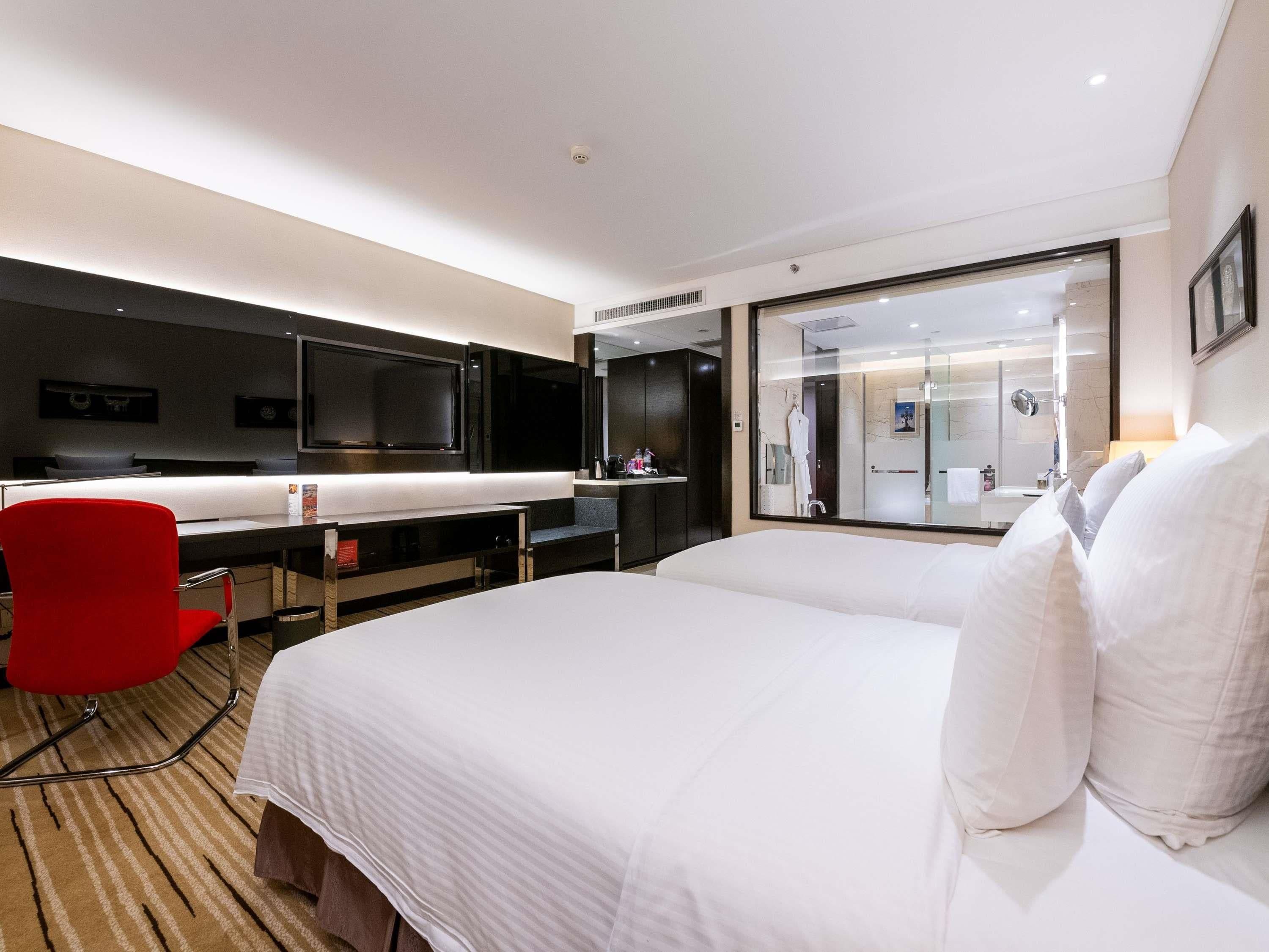 A modern hotel room with a large bed, a red chair, a desk with a flat-screen TV, and a bathroom visible through a glass partition.